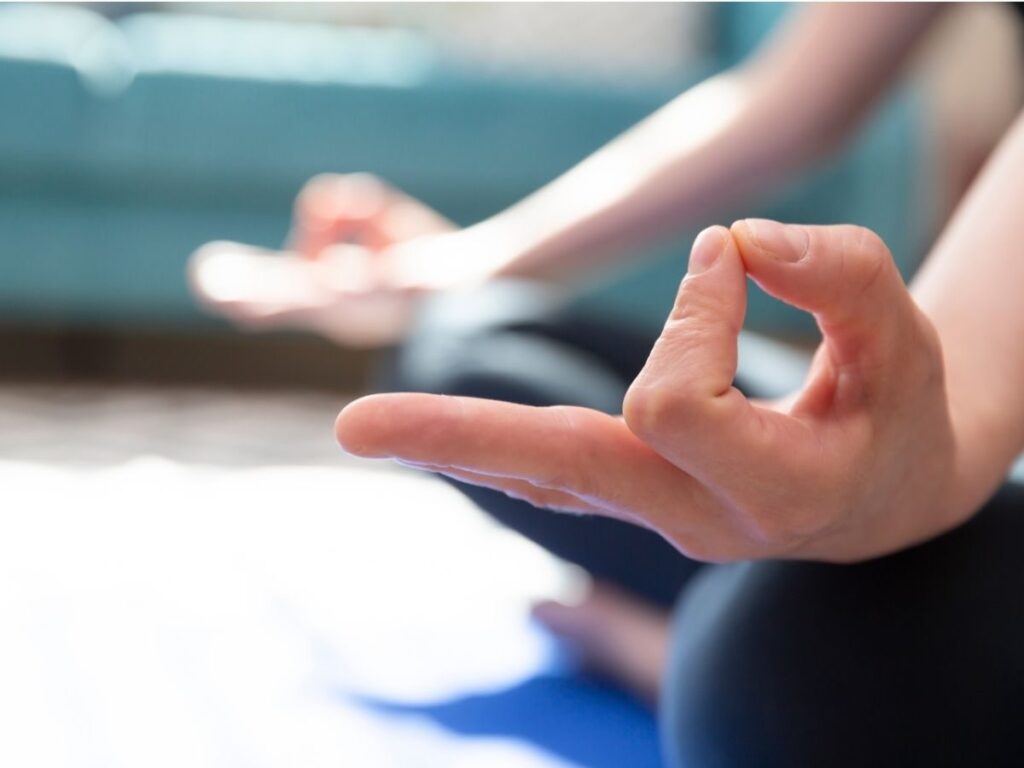 Gros plan sur deux mains en posture de méditation, les doigts formant le Gyan mudra. La personne est assise sur un tapis, vêtue de vêtements sombres. Un canapé flou apparaît en arrière-plan. L'atmosphère paraît calme et paisible.