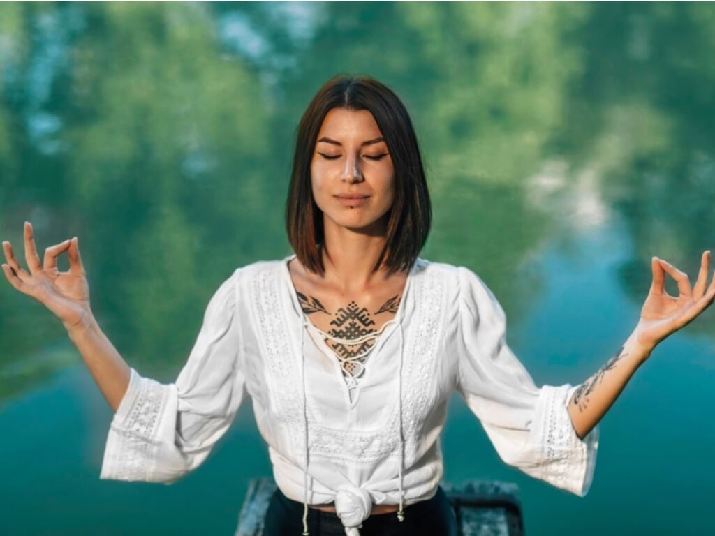 Une personne aux longs cheveux noirs médite en plein air, debout, les yeux fermés et les bras levés, formant des cercles avec les doigts. Elle se trouve près d'une eau calme, d'un bleu verdâtre, où se reflètent des plantes vertes. Elle porte un haut blanc avec des tatouages visibles.