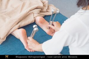 Une personne est allongée sur une surface bleue avec une serviette beige couvrant le haut de son corps. Une autre personne utilise deux diapasons métalliques sur la plante des pieds de la personne. Le texte en bas de la page indique : « Diapason thérapeutique : tout savoir avec Meozen.