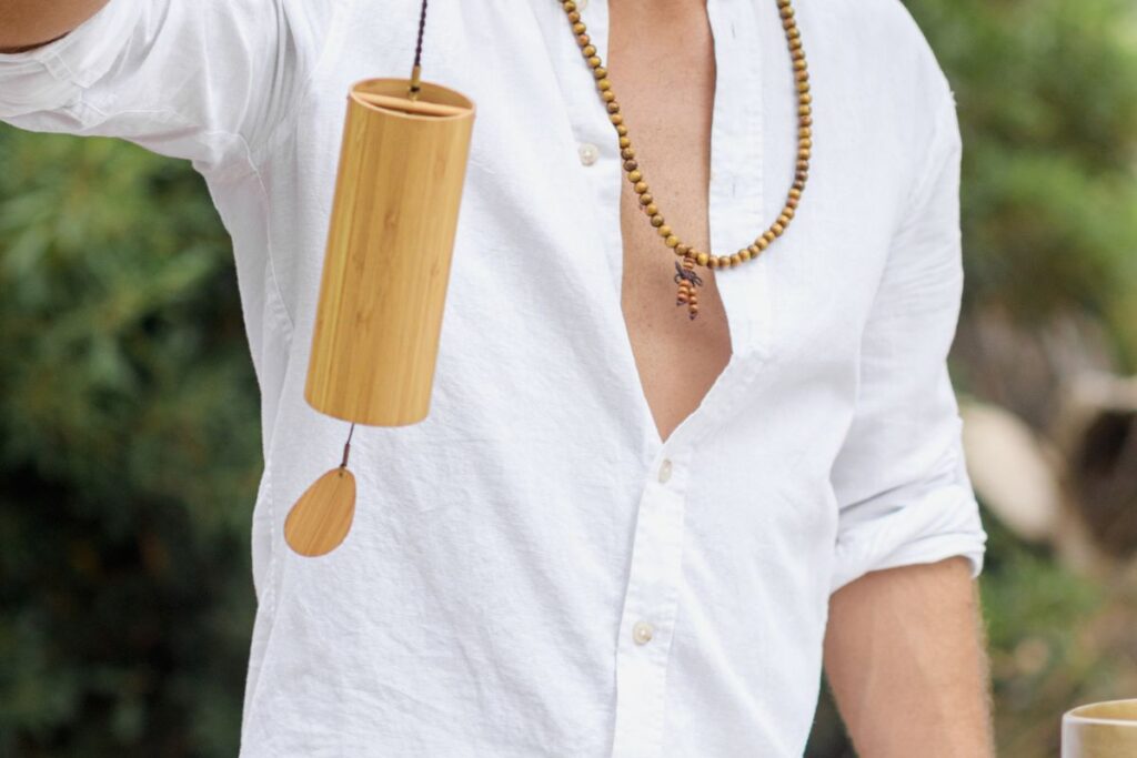 Une personne portant une chemise blanche et un collier de perles tient un carillon éolien cylindrique en bambou. Le carillon est doté d'une petite pièce en bois en forme de feuille suspendue en dessous. L'arrière-plan est une verdure floue.