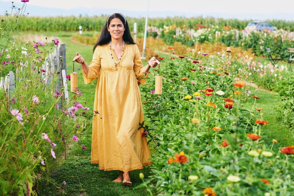 Une femme vêtue d'une longue robe jaune marche pieds nus dans un jardin fleuri aux couleurs vives. Elle tient deux ficelles attachées à des poteaux en bois. Le jardin est luxuriant, avec des fleurs aux couleurs variées, sur fond de champs et de ciel dégagé.