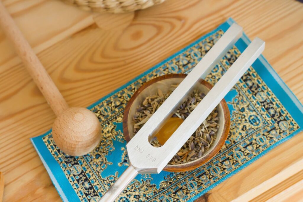 Un diapason repose sur une surface en bois à côté d'un petit bol rempli d'herbes et d'un cristal jaune. Un maillet en bois se trouve à proximité, le tout posé sur un tissu à motifs bleus.