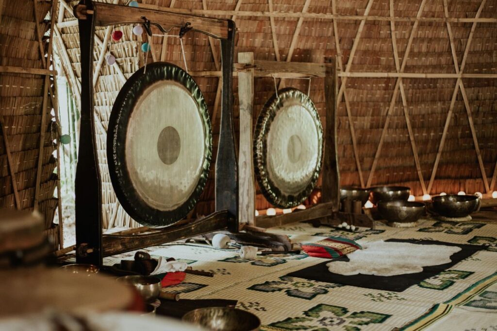 Une pièce avec deux grands gongs montés sur des cadres en bois est présentée. La pièce est dotée d'un mur en chaume et d'un tapis à motifs sur le sol, ainsi que de divers petits bols en métal disposés autour de l'espace.