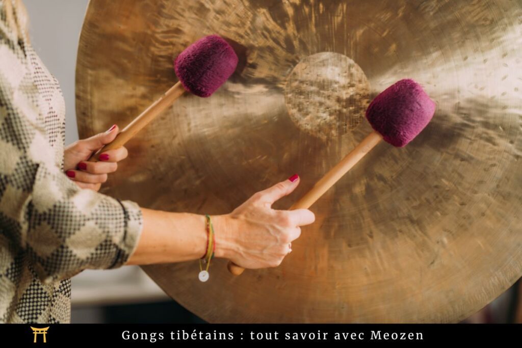 Un homme tient deux maillets et frappe un grand gong. Les maillets ont des têtes violettes et les mains de l'homme sont visibles. Le gong a une surface circulaire brillante avec un motif rayonnant à partir du centre. Le texte en bas mentionne "Gongs tibétains".