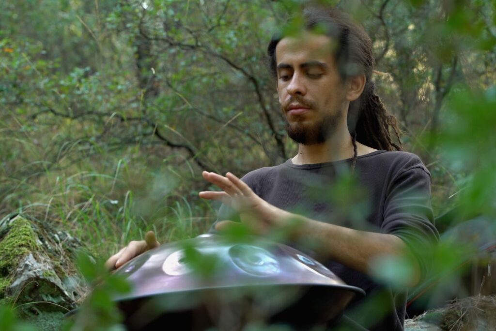 Une personne aux cheveux longs et à la barbe est assise à l'extérieur dans une zone boisée, jouant du handpan. Le feuillage environnant est vert et luxuriant, et la personne semble concentrée sur l'instrument.