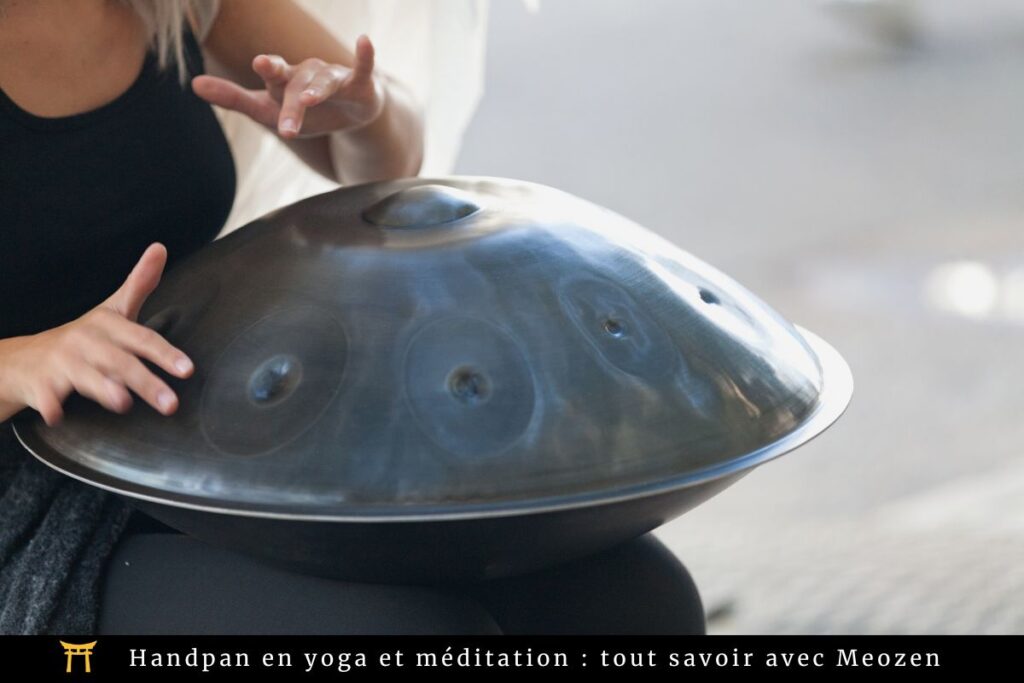 Une personne joue du handpan avec ses doigts. L'instrument est métallique et présente plusieurs zones alvéolées. L'arrière-plan est flou. Le texte en bas indique : « Handpan en yoga et méditation : tout savoir avec Meozen ».