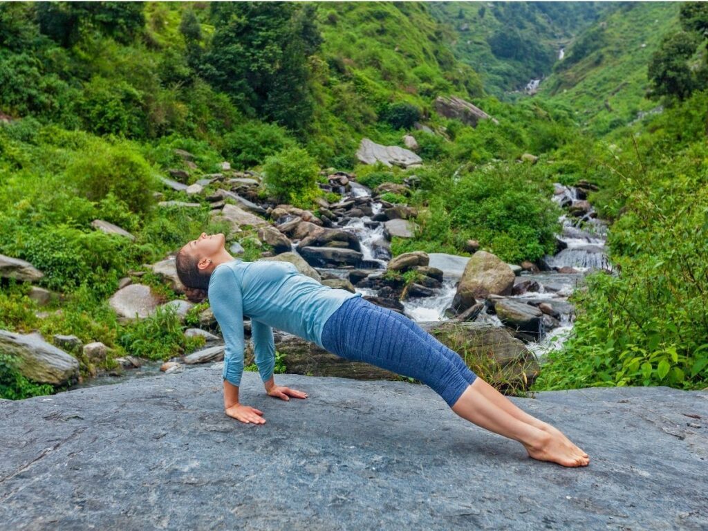 Une personne effectue une planche ascendante sur un gros rocher dans une région montagneuse verdoyante. Une petite cascade et un ruisseau sont visibles en arrière-plan. La personne porte un haut bleu clair et un pantalon bleu foncé.