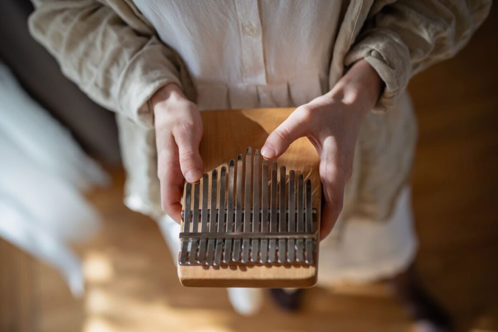 Une personne tient un kalimba, un instrument de musique en bois avec des lames en métal, sur un fond flou. La personne porte une chemise et une veste de couleur claire.