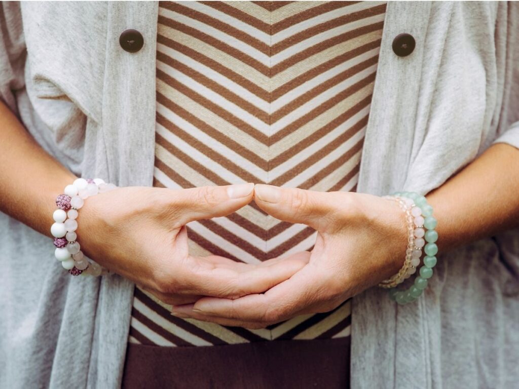 Une personne vêtue d'une chemise rayée et d'un cardigan gris place ses mains sur son ventre dans un geste détendu et symétrique. Elle porte des bracelets de perles blanches et vertes à chaque poignet.