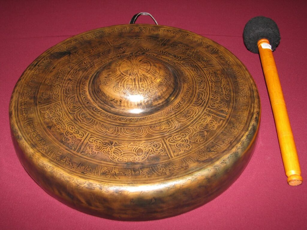 Un handpan en bronze aux motifs gravés complexes repose sur une surface marron. À droite, un maillet orange avec une extrémité de frappe noire et arrondie est placé parallèlement à l'instrument.