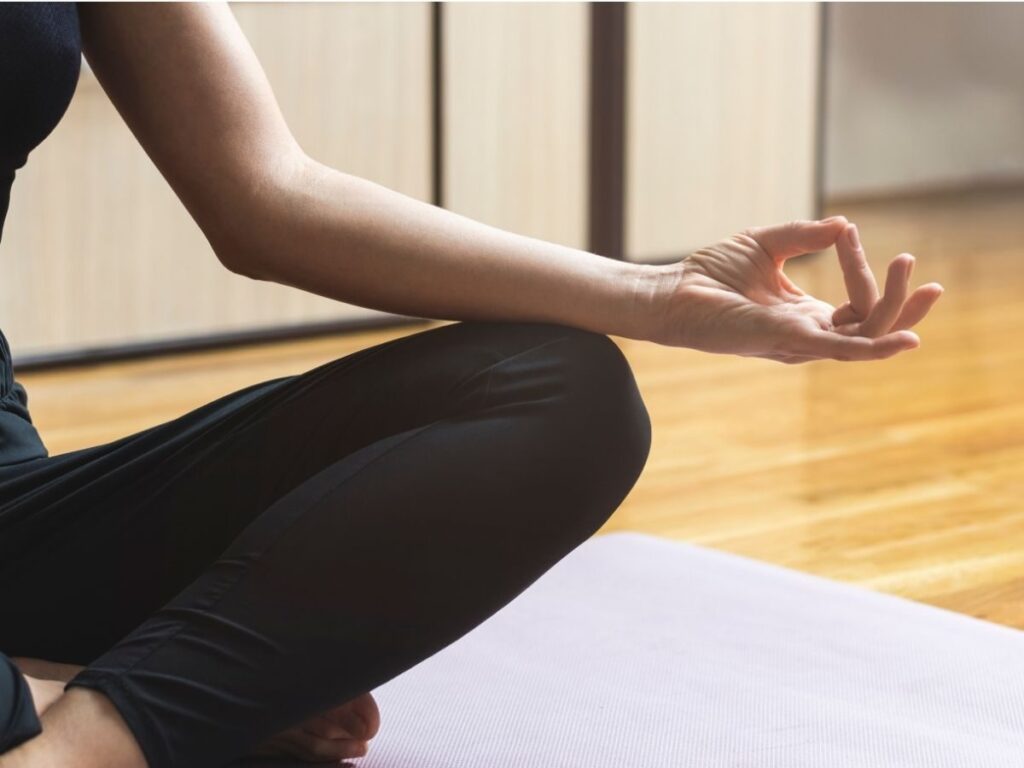 Une personne est assise en tailleur sur un tapis de yoga violet clair, vêtue d'un legging noir. Sa main droite repose sur son genou, le pouce et l'index se touchant, formant un mudra. Le sol est en bois et la lumière filtre doucement.