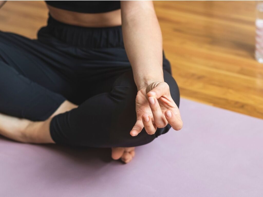Une personne est assise en tailleur sur un tapis de yoga violet. Elle porte une tenue de sport noire et pose une main sur son genou, les doigts en mudra. Le parquet et une partie d'une bouteille d'eau sont visibles en arrière-plan.