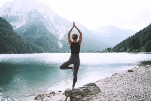 Une personne se tient sur une jambe dans une pose de yoga sur le rivage rocheux d'un lac, face à un lac calme entouré de montagnes boisées sous un ciel nuageux.
