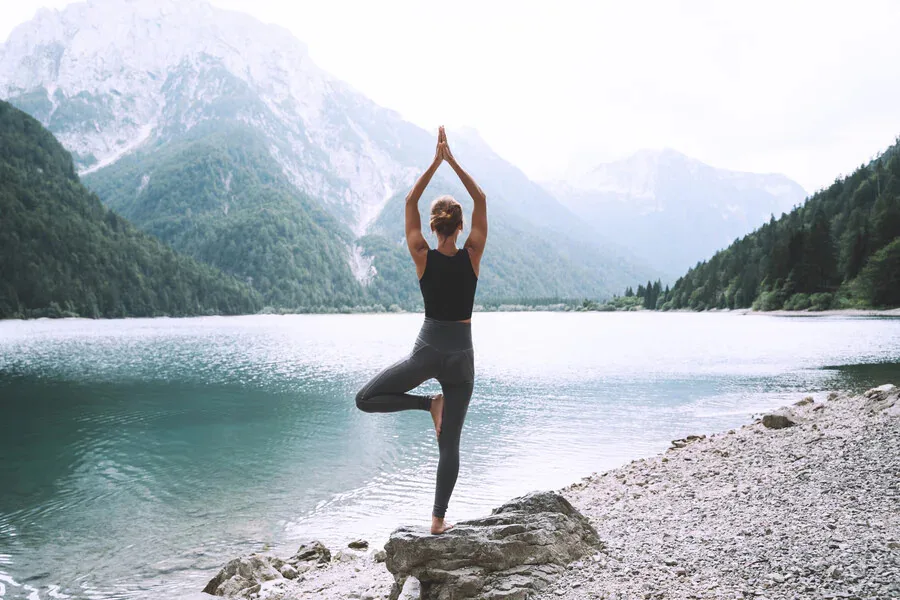 Une personne se tient sur une jambe dans une pose de yoga sur le rivage rocheux d'un lac, face à un lac calme entouré de montagnes boisées sous un ciel nuageux.